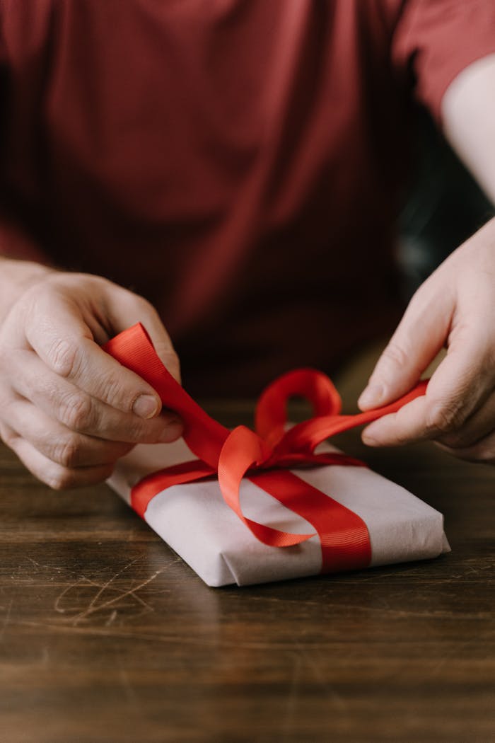 Close-up of hands tying a red ribbon on a gift box on a wooden surface.